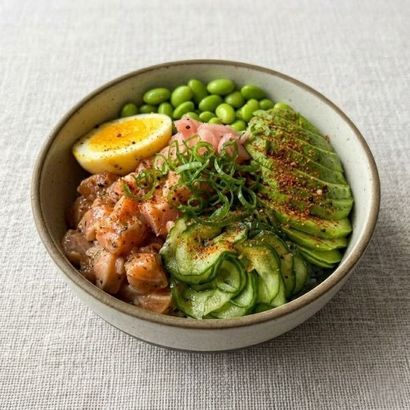 Bowl of salmon poke with avocado, cucumber, edamame, and lemon wedge on a light tablecloth