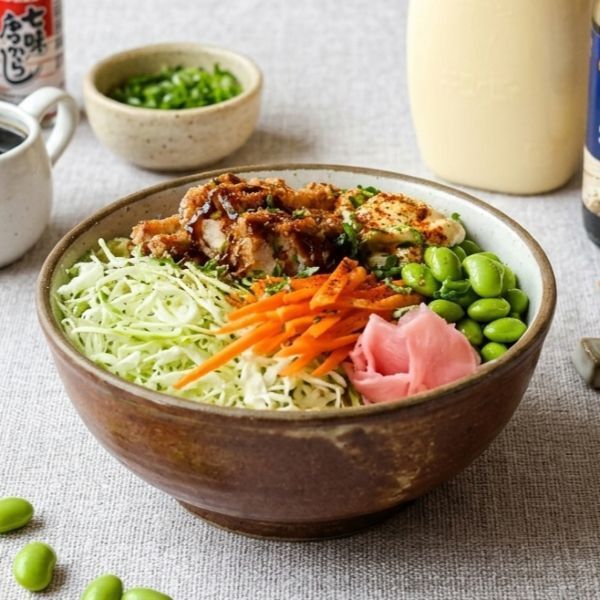 Top-down view of two poke bowls, soy sauce, mayonnaise, pickled ginger, green onions, and chopsticks on a textured cloth.