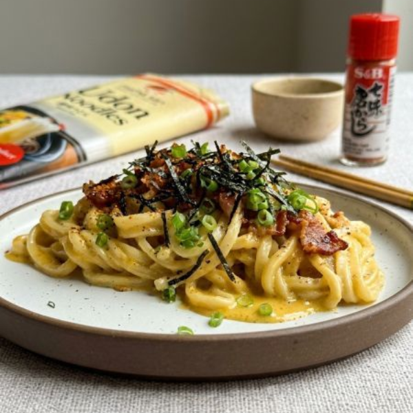 Two plates of udon noodles with creamy sauce and toppings, alongside a package of udon noodles, chopsticks, and spices.