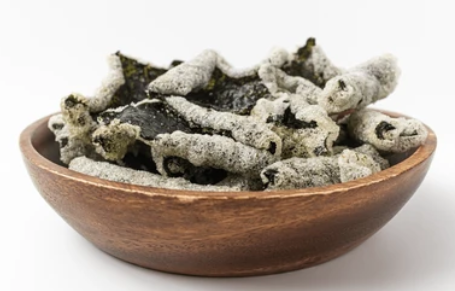 Bowl of crispy, green seaweed snacks coated in white powder, on a white background.