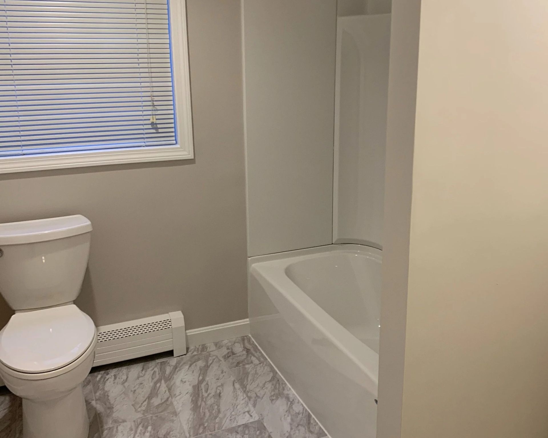 Bathroom with toilet, bathtub, and window with blinds. Gray walls, light gray floor, and white fixtures.