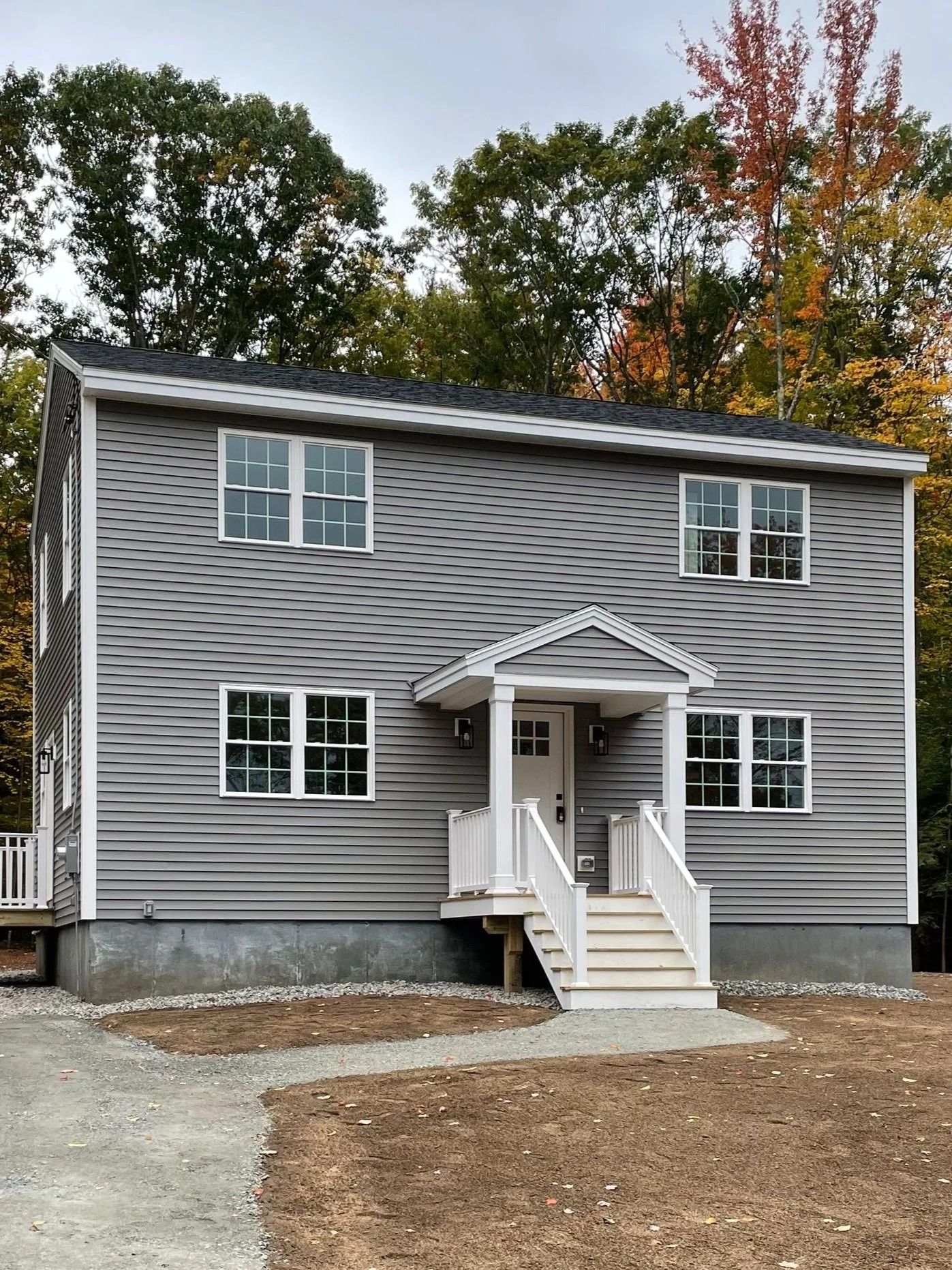 Gray two-story house with white trim, porch, and windows. Brown trees and cloudy sky in background.
