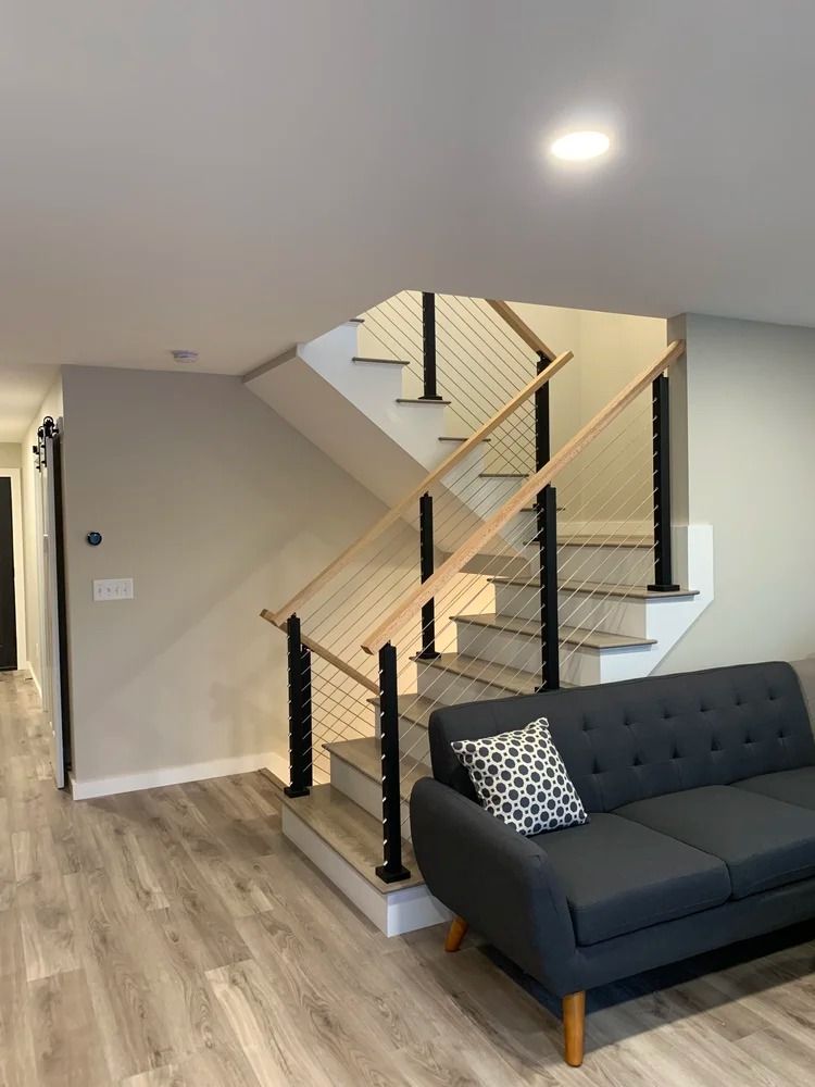 Modern staircase with light wood handrails, black posts, and cable railing. Gray walls, light flooring, and a gray sofa.