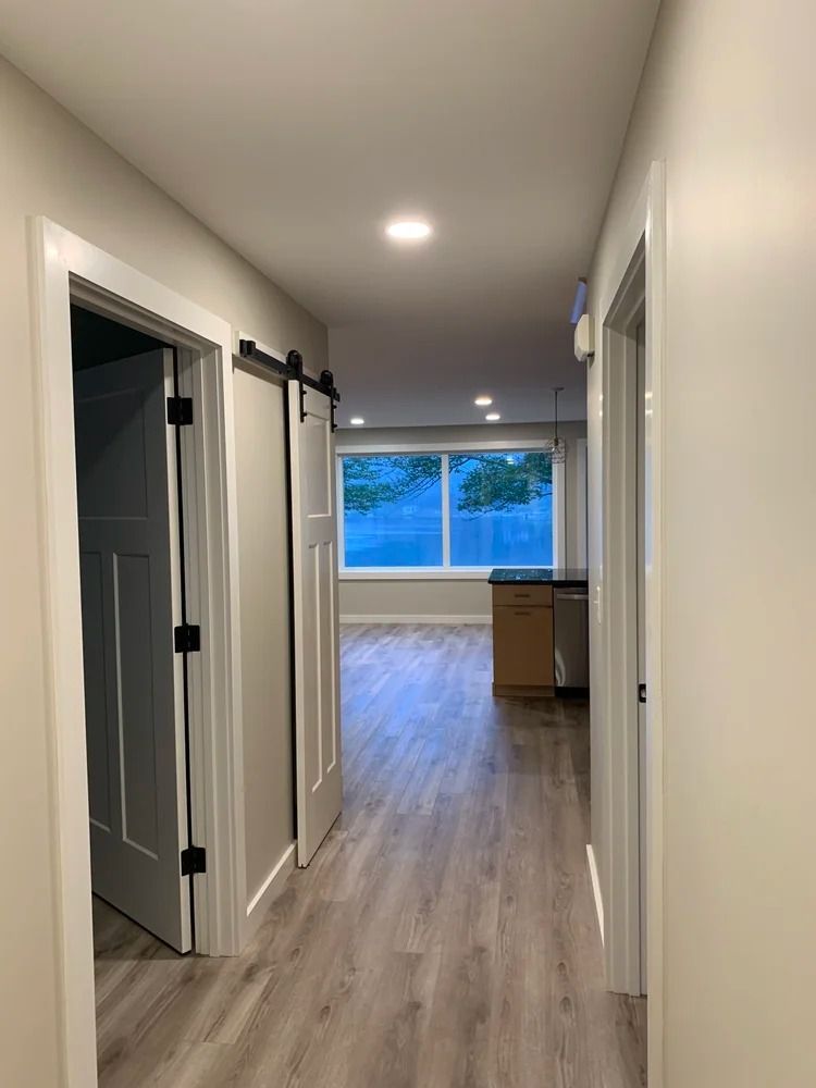 Hallway with wood floors, two white doors, one with a sliding barn door style. View to a kitchen.