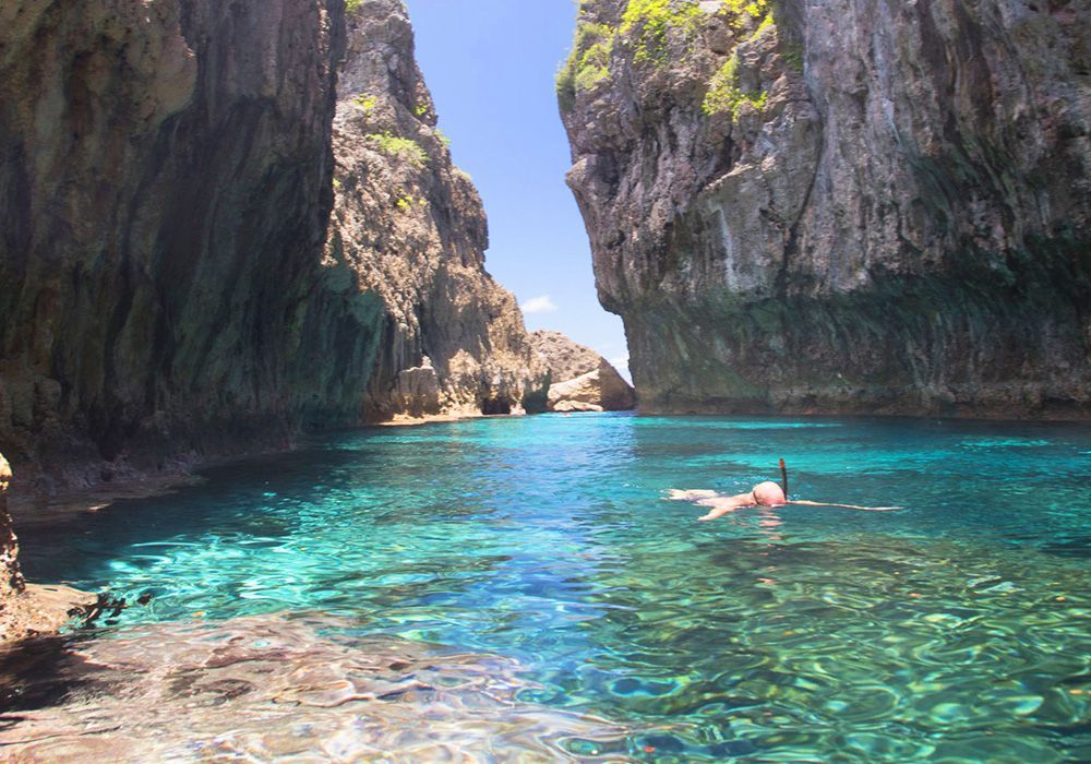 Woman snorkeling in clear turquoise water between rocky cliffs.