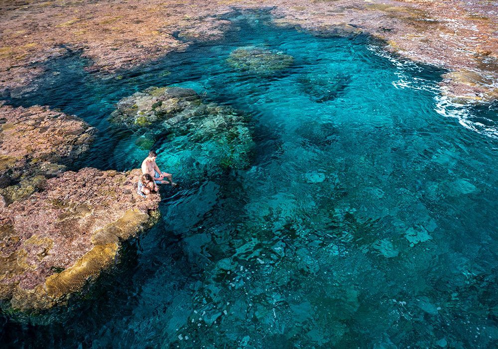 Woman sits on rocky shore, overlooking clear turquoise ocean.