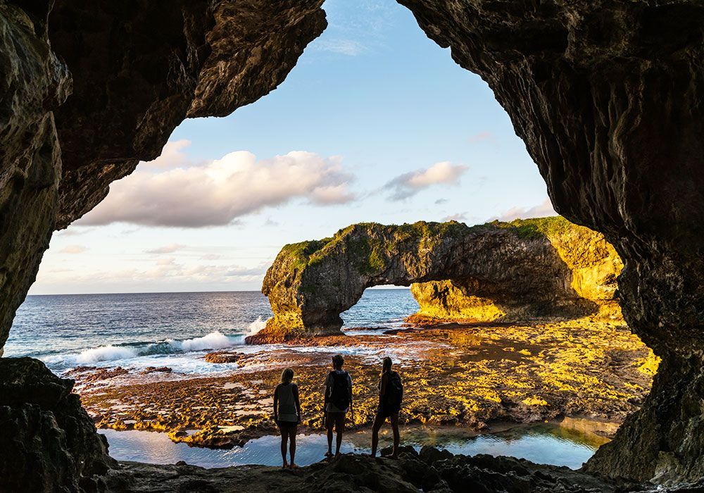 Three people stand in a cave, looking out at a natural arch over the ocean.