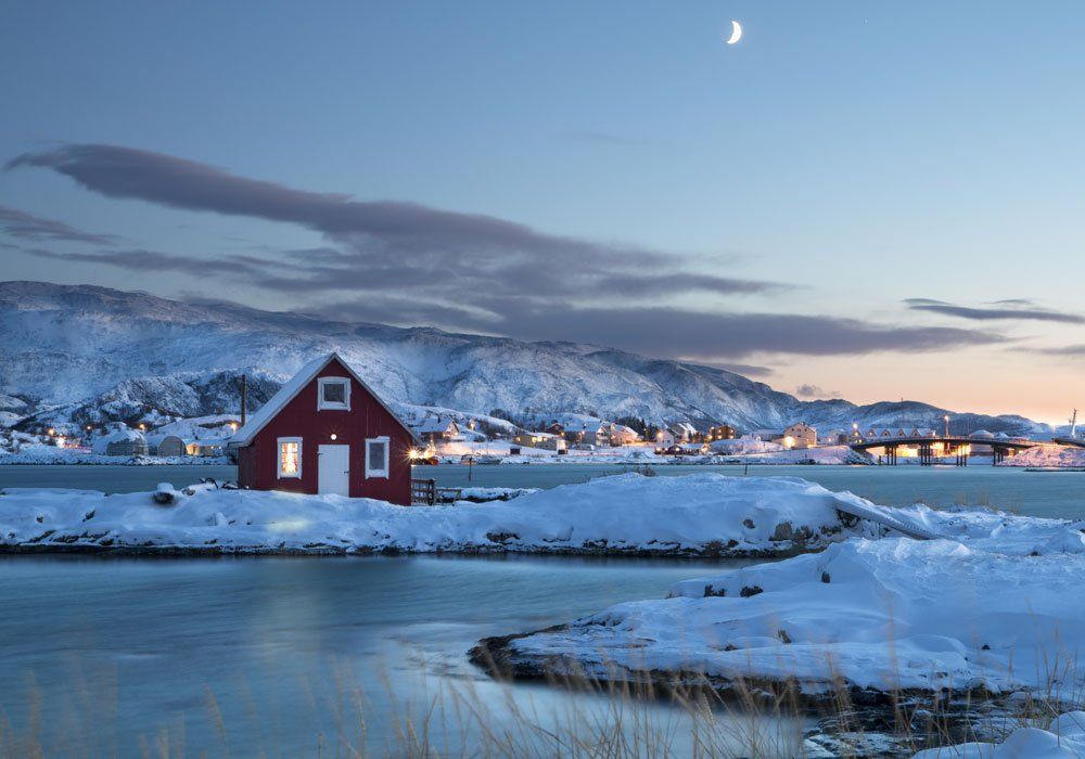 A red house is sitting on a snow covered island next to a body of water.