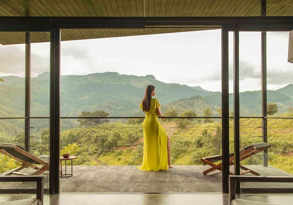 A woman in a yellow dress is standing on a balcony overlooking mountains.
