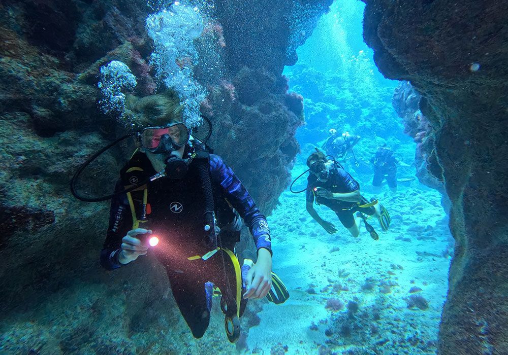 Divers explore an underwater cave, one holding a light, in a vibrant blue ocean.