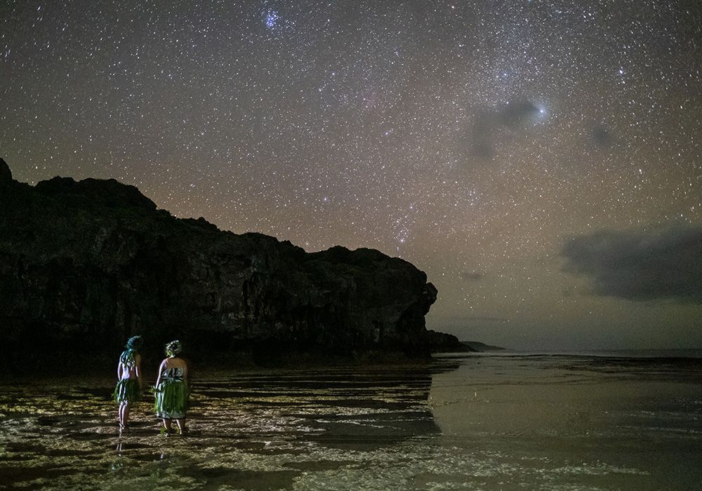 Two people stand on a rocky shore, gazing at a starry night sky over the ocean.