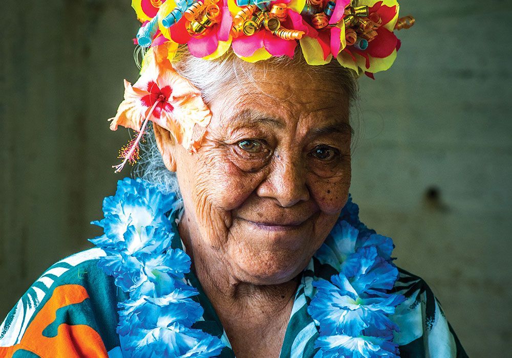 Elderly woman wearing floral crown and lei smiles.