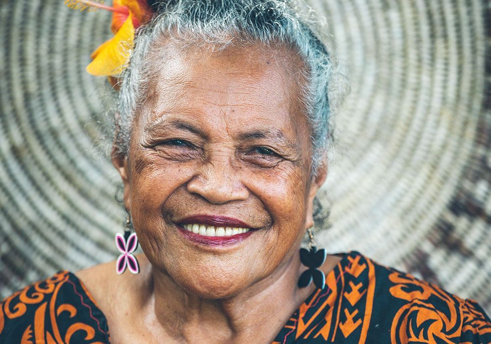 Smiling older woman with gray hair, flower in her hair, patterned earrings, and colorful shirt.