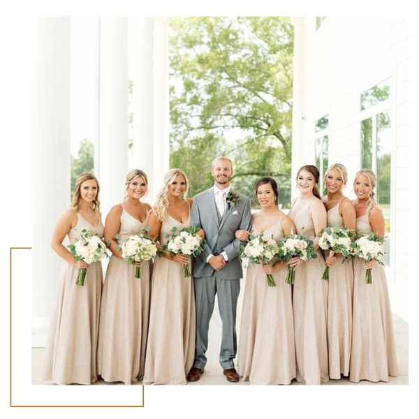 Wedding party posing outdoors in white columns, with a groom in gray and bridesmaids in blush dresses holding bouquets