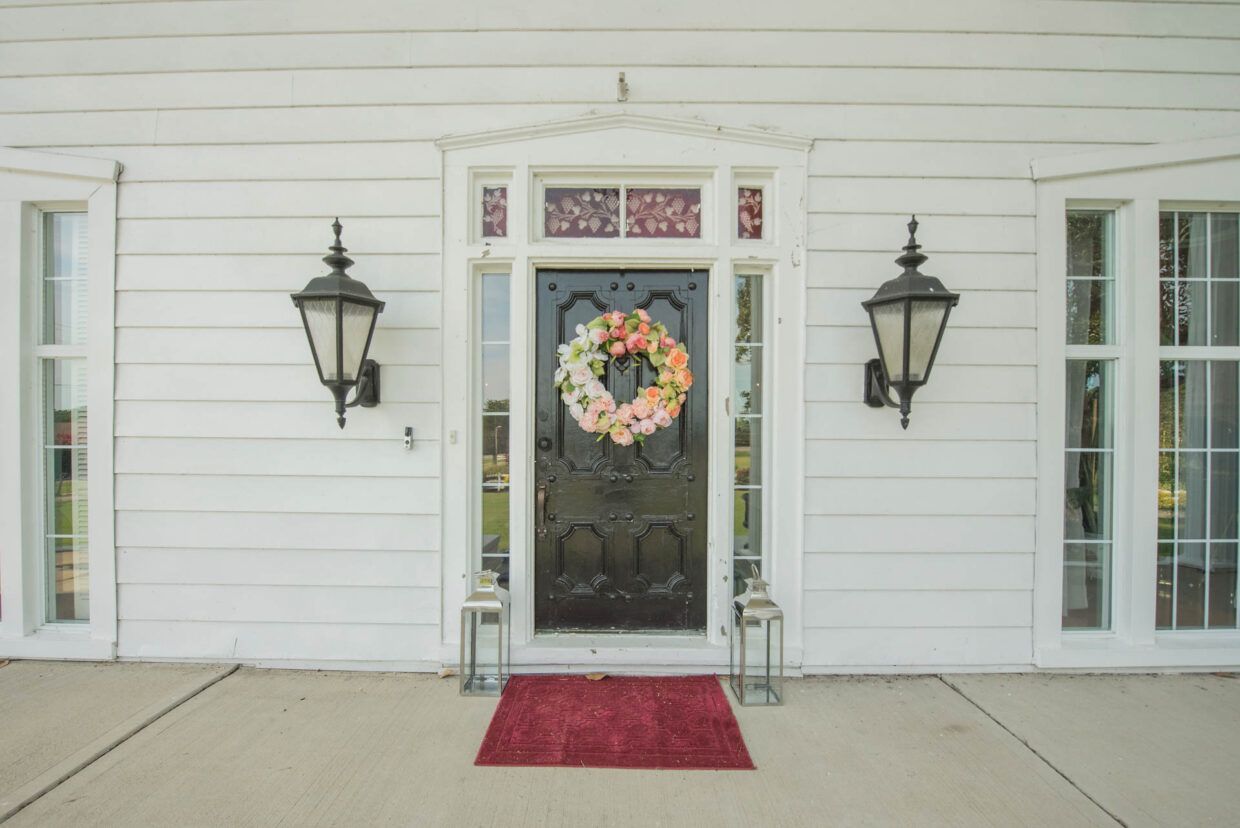 White house entrance with a black door, floral wreath, and lanterns. A red rug and windows frame the door.