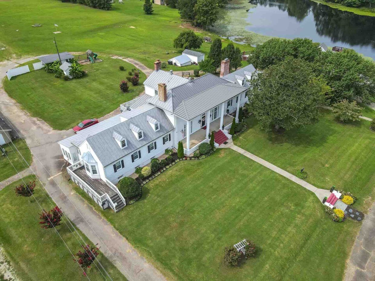 Aerial view of a white mansion with columns, surrounded by green lawns, a lake, and trees.