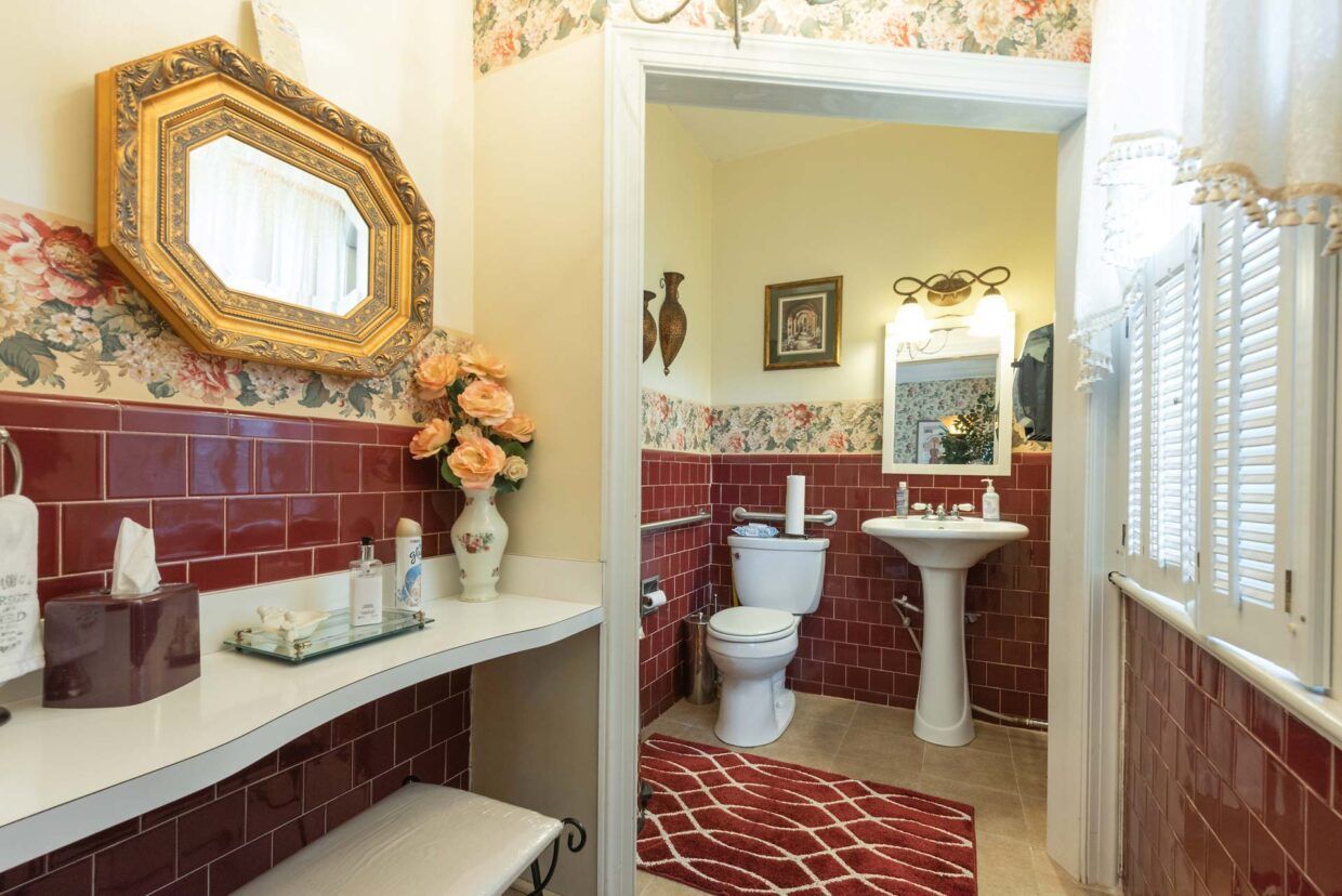 Bathroom with burgundy tile, floral wallpaper, vanity, toilet, sink, and ornate mirror.