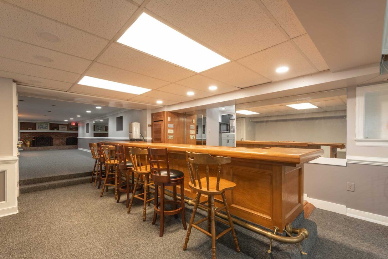 Basement bar with wooden counter, stools, and recessed lighting. Gray carpet, neutral walls.