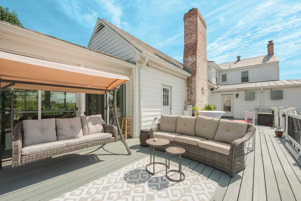 Outdoor deck with seating: swing, sofa, and small tables, against a white house with a brick chimney.