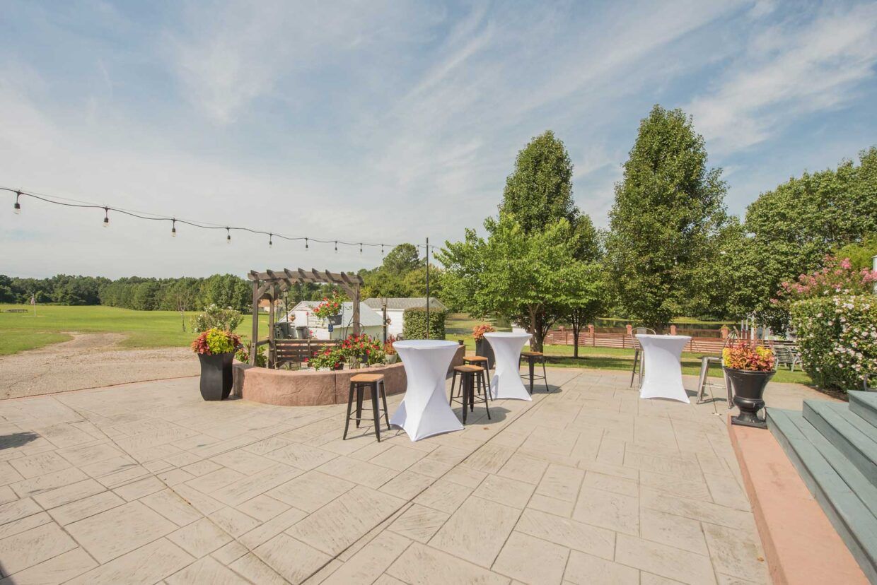 Outdoor patio with cocktail tables, potted flowers, and string lights, overlooking a grassy field.