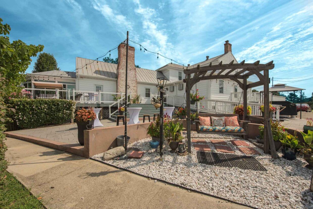 Courtyard with white house, pergola, string lights, and seating area on a sunny day.