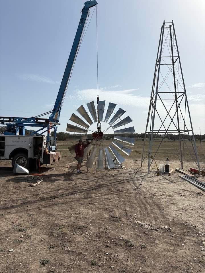 A crane lifts a large metal windmill wheel to be attached to a tall, steel lattice tower on a rural, dry landscape.