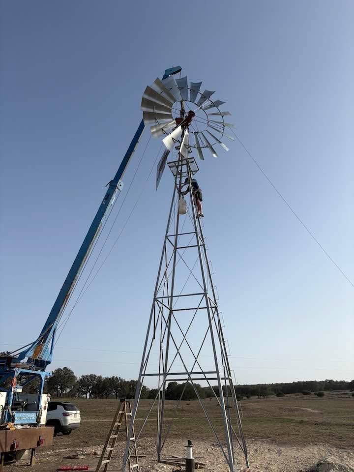 A worker on a tall windmill tower uses a crane to assist with maintenance on the blades against a clear sky.