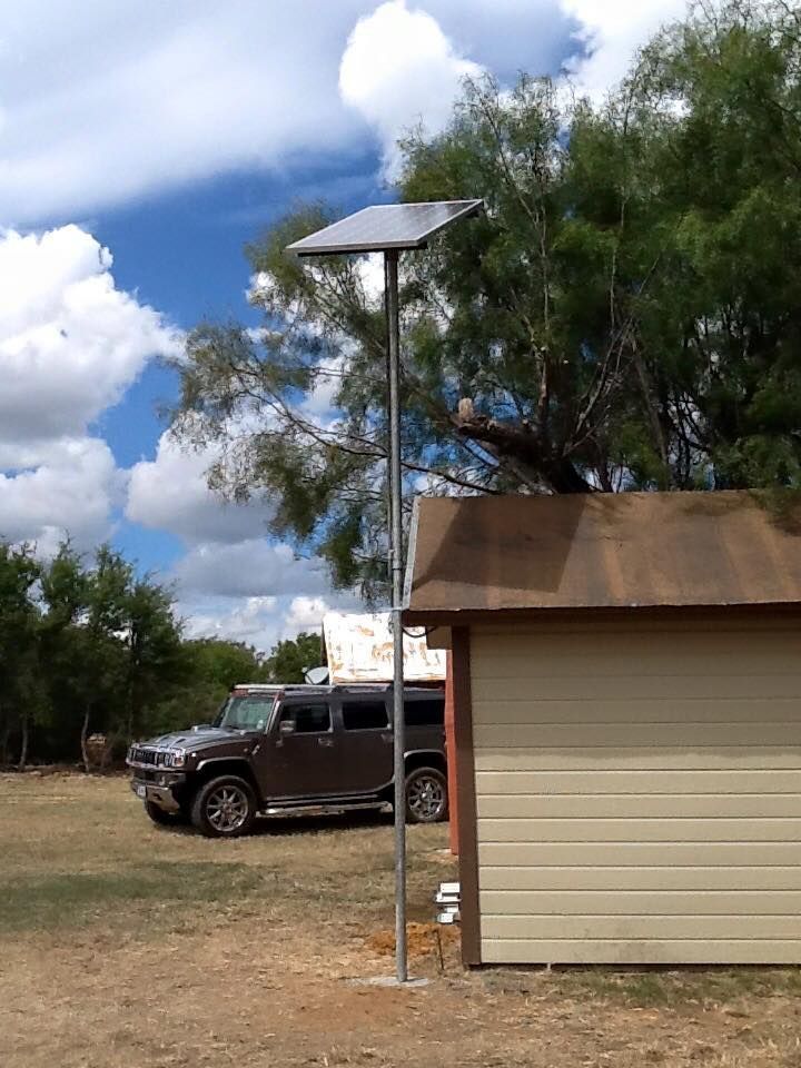 A solar panel mounted on a tall pole beside a shed, with a brown SUV parked in the background under a cloudy sky.