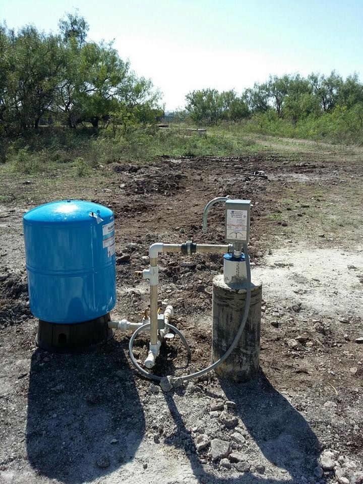 A blue pressurized water tank connected to a well pump housing in a sunny, rural outdoor field.