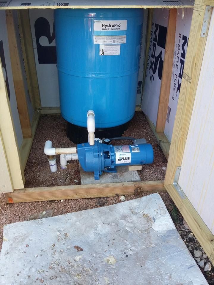 A blue water pressure tank and pump system inside a wooden, insulated utility enclosure.