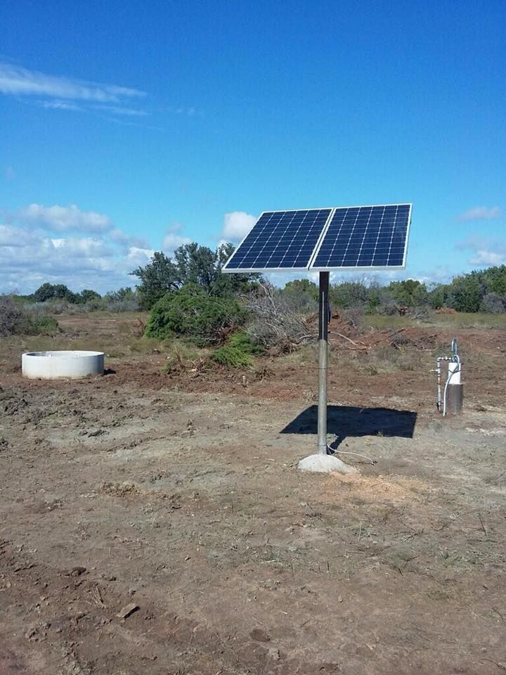 A solar panel array on a metal pole in a dry, open field with a round concrete basin nearby under a clear blue sky.