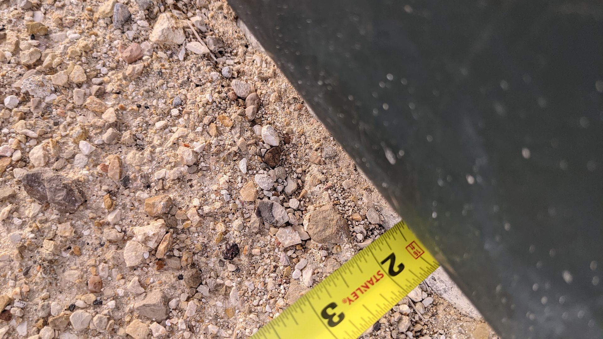 A yellow tape measure placed against the edge of a dark object on a gravel surface, showing a measurement of 2 inches.