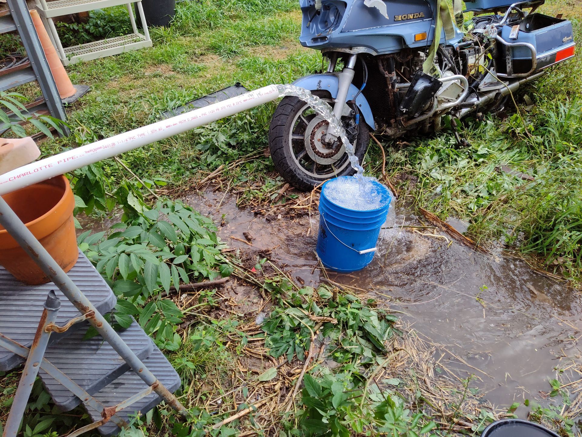 Water flows from a white PVC pipe into a blue bucket parked next to a blue motorcycle in a grassy outdoor area.