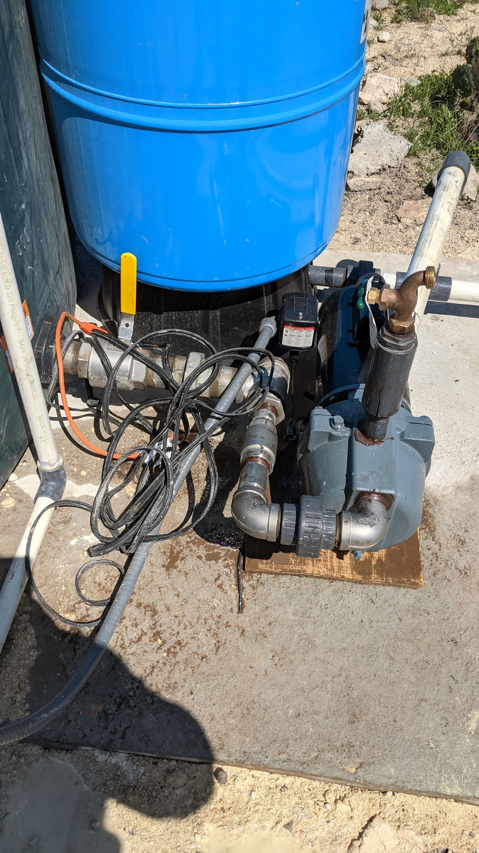 A blue pressure tank connected to a gray water pump on a concrete pad with visible piping and electrical wiring.