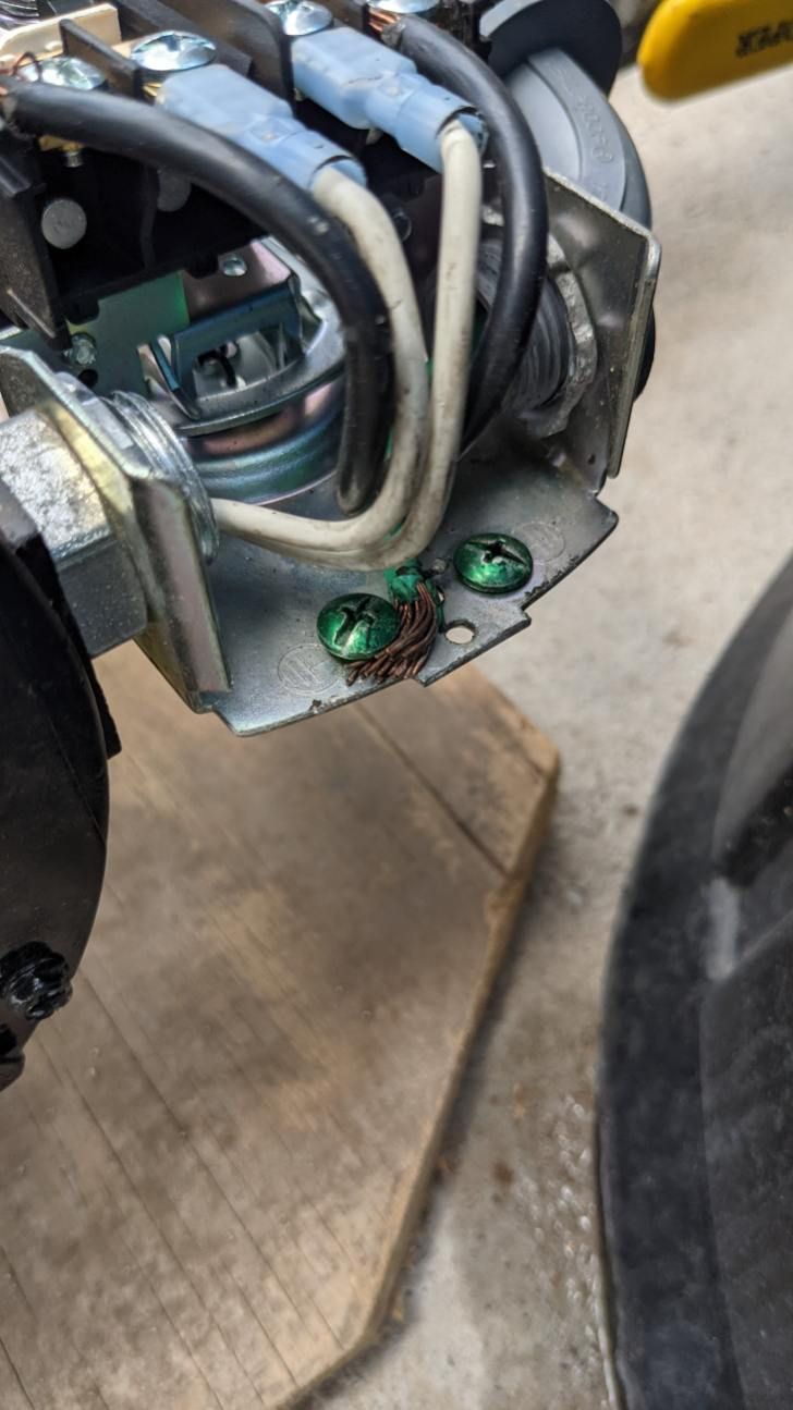 Close-up of internal electrical wiring connections on a metal component with green grounding screws on a wood surface.