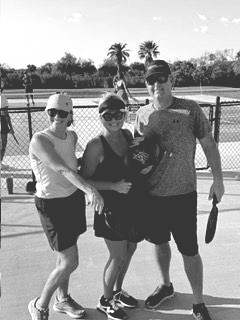 Three people posing with tennis rackets on an outdoor tennis court, with nets and palm trees behind them.