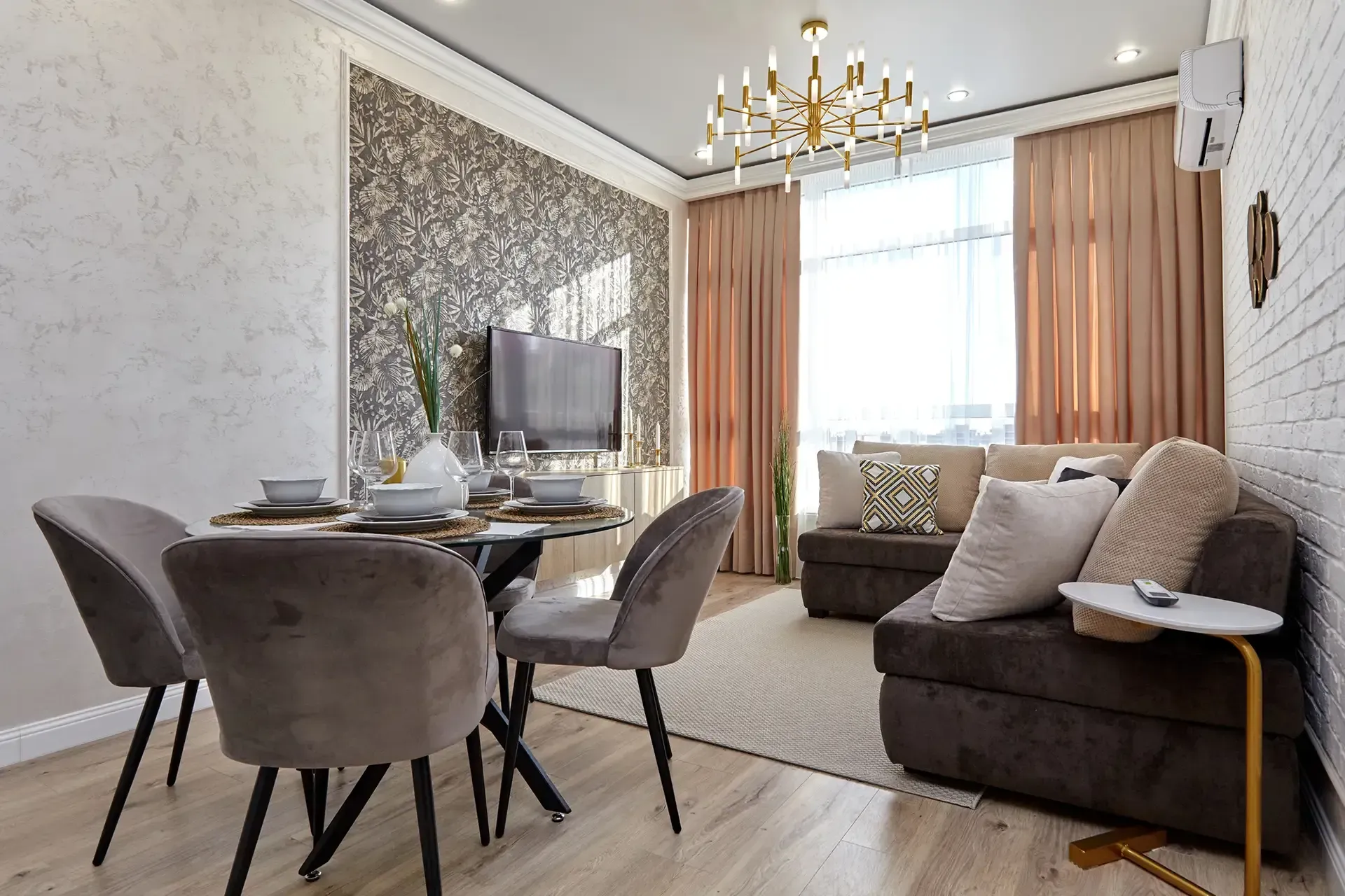 Living room with dining table, sofa, and large window with curtains; gold chandelier and neutral color scheme.