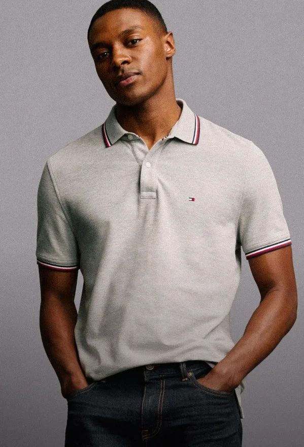 A man wearing a grey polo shirt and jeans is standing with his hands in his pockets.