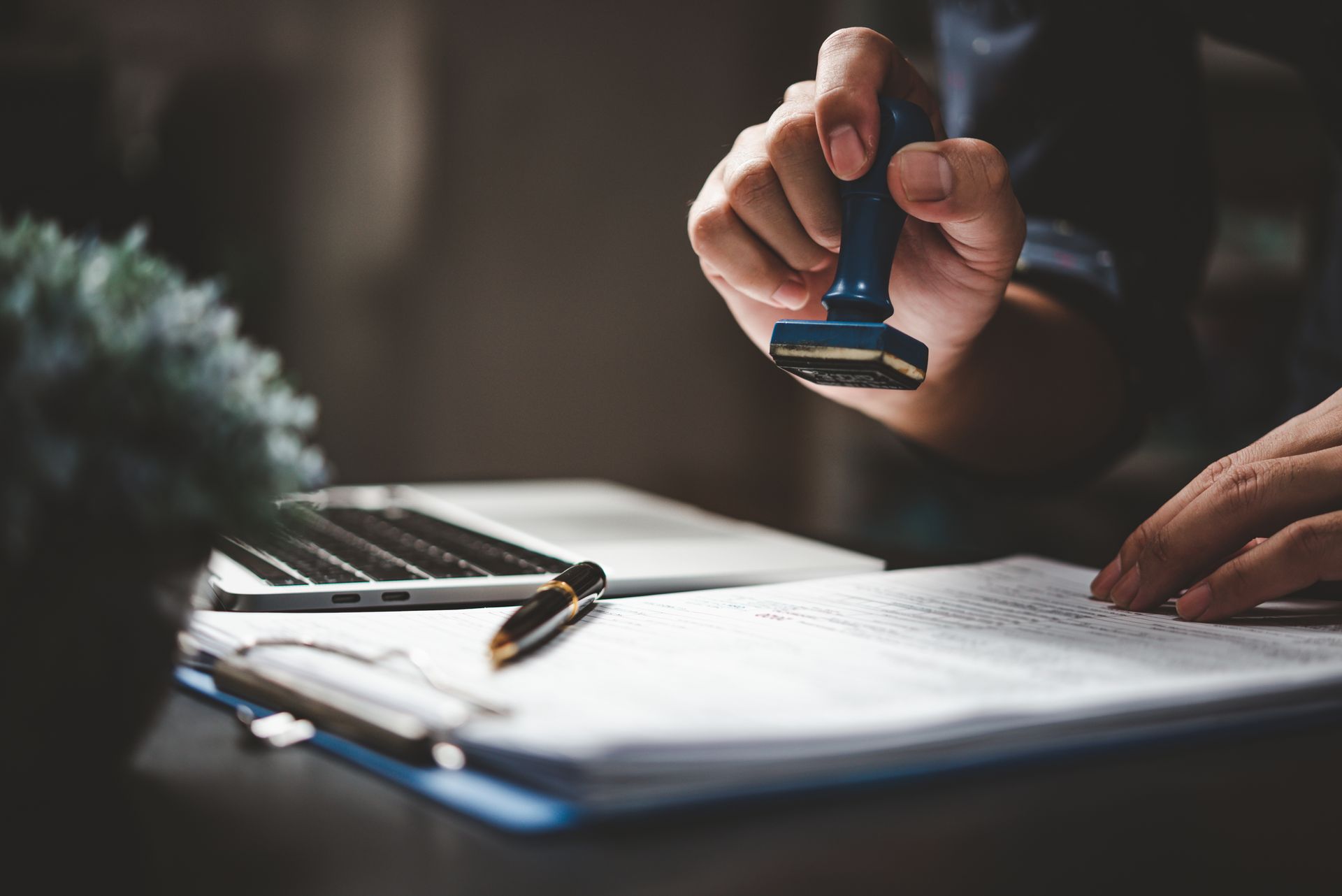 Person stamping documents on a desk beside a laptop and pen in a dim office setting.