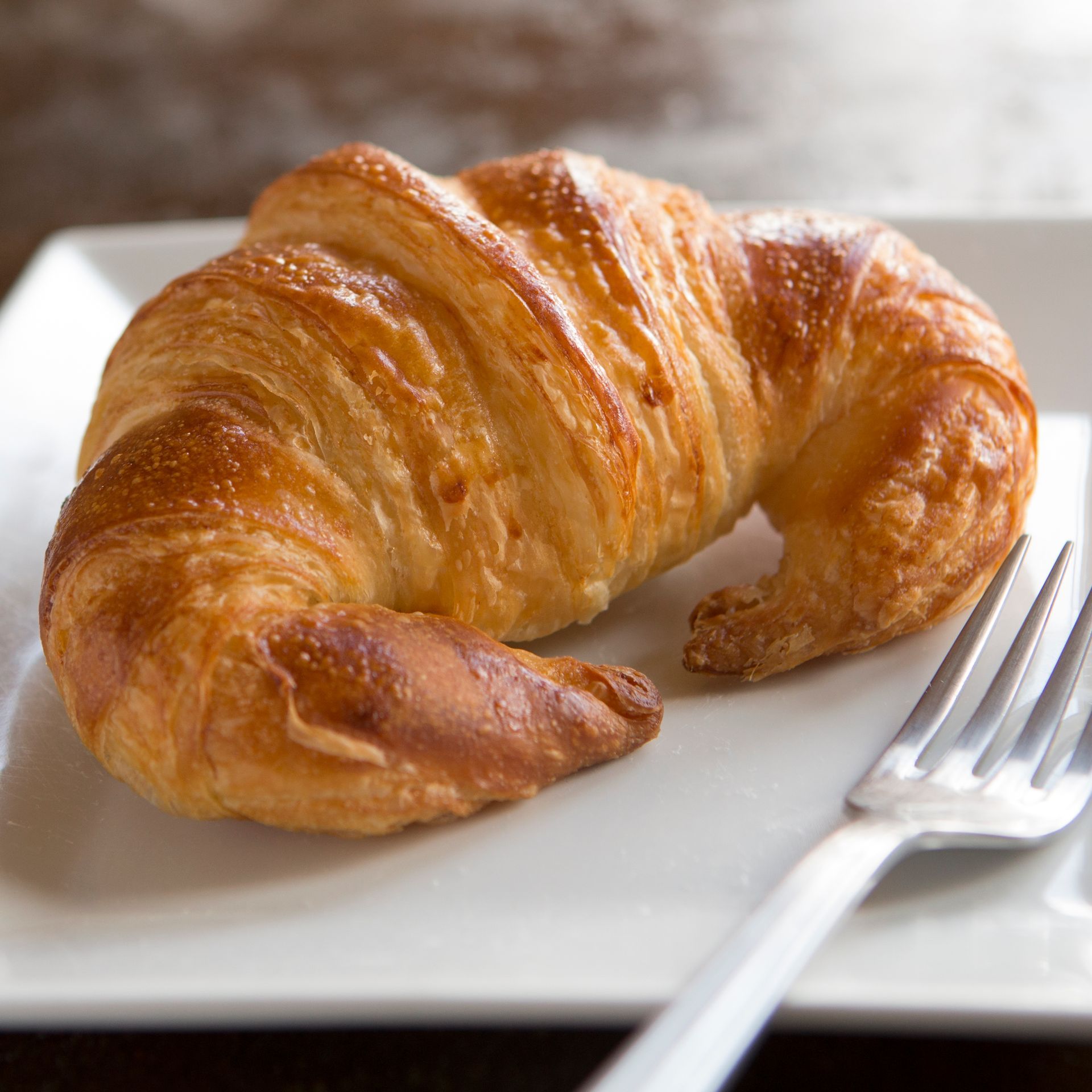 A croissant is sitting on a white plate next to a fork.