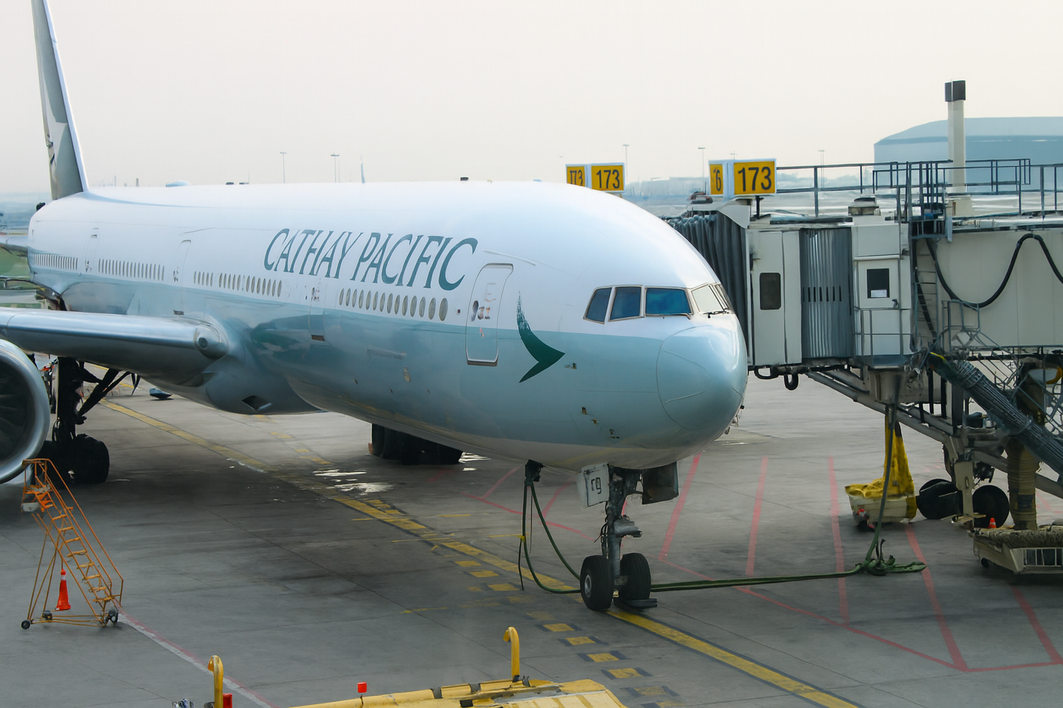 Cathay Pacific airplane at a gate. White fuselage with green logo, connected to a jet bridge.