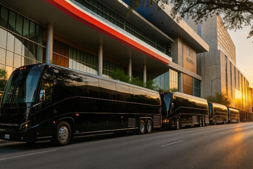 Four black charter buses parked along a street in front of a modern building; golden sunset.