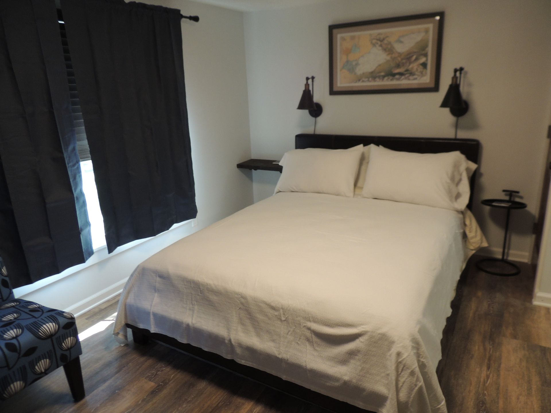 Bright bedroom with a queen bed, dark curtains, wall art, and wood floors
