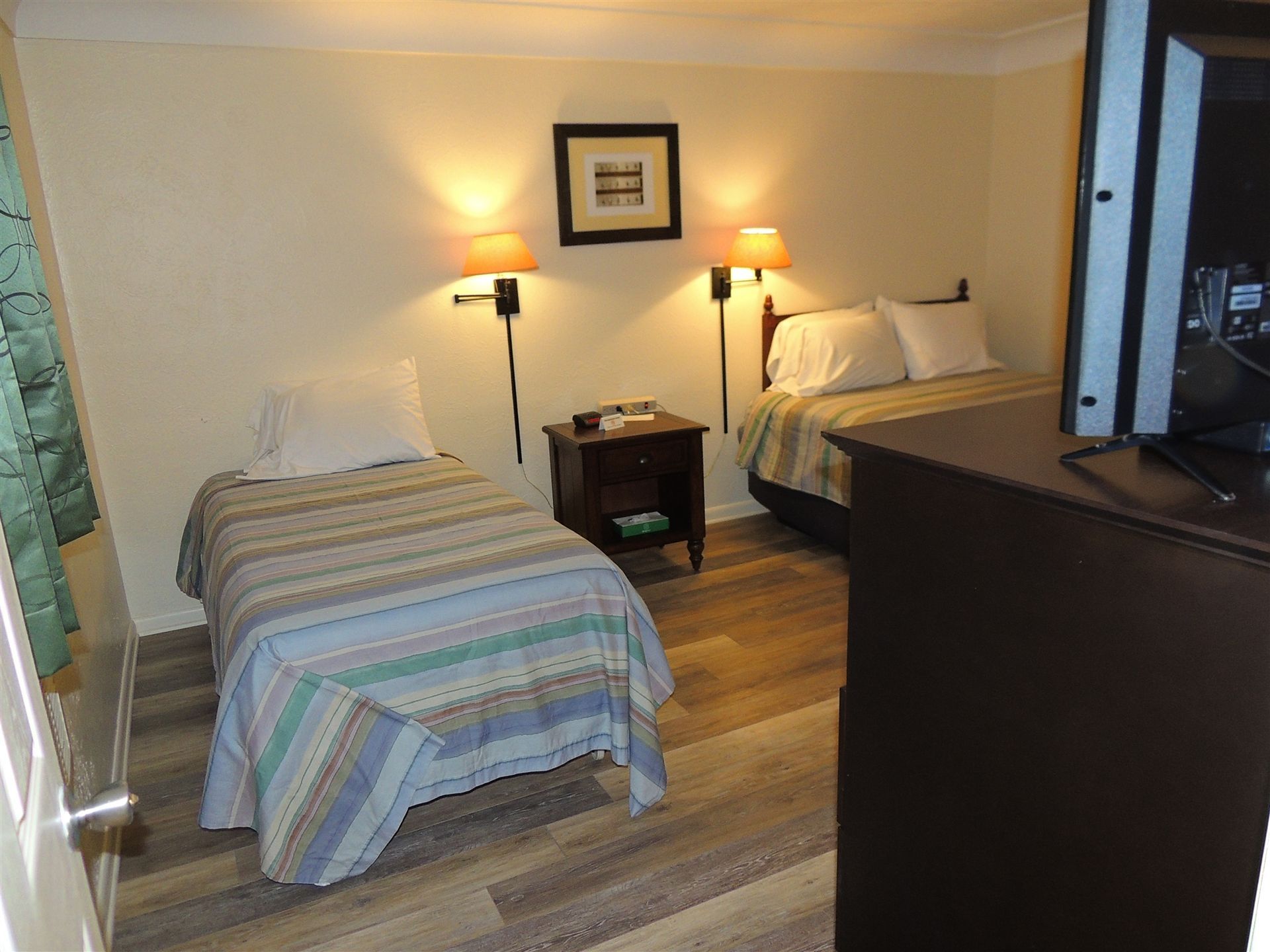 Small bedroom with two twin beds, bedside table, lamps, and a dark dresser in the foreground