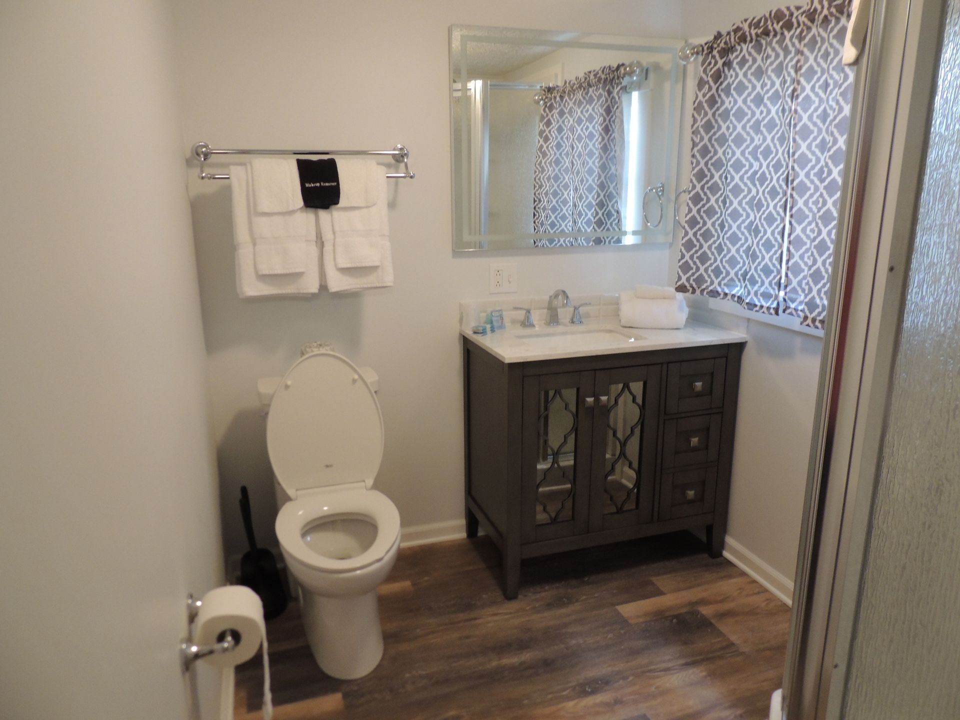 Bathroom with toilet, vanity sink, mirror, and window with patterned curtains on wood-look flooring
