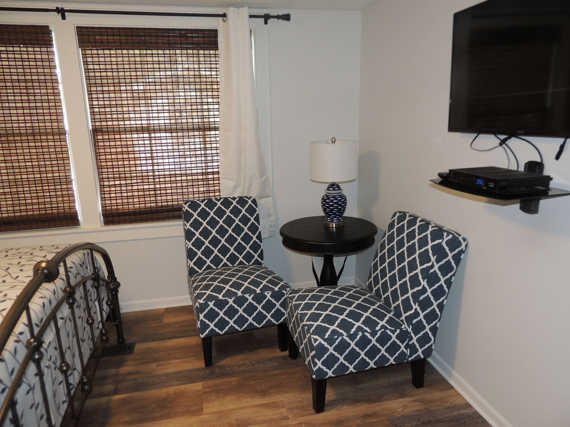 Small bedroom with two patterned chairs, a round table, wall TV, and windows with bamboo blinds