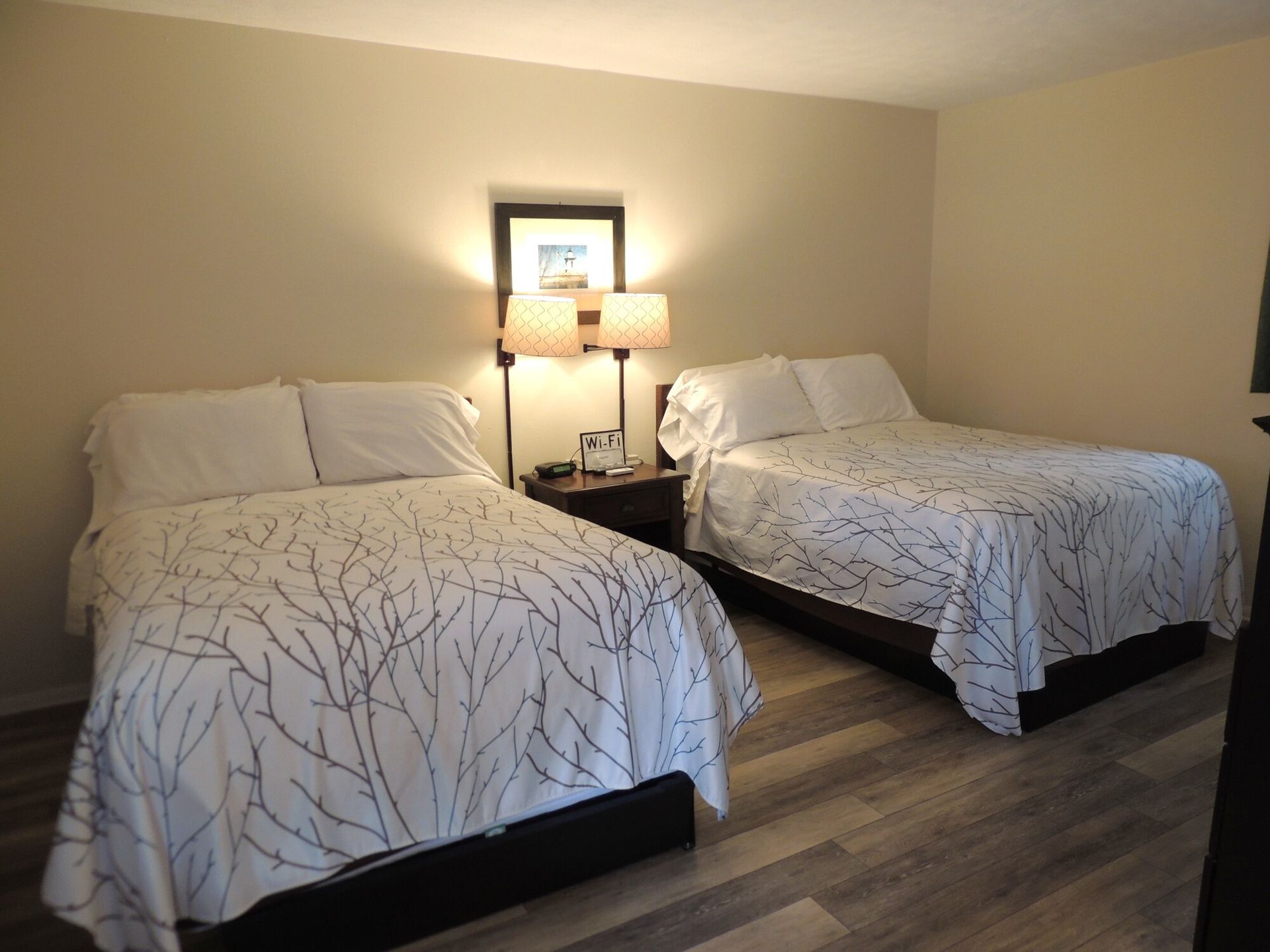 Two neatly made beds in a beige hotel room with a bedside lamp and window.