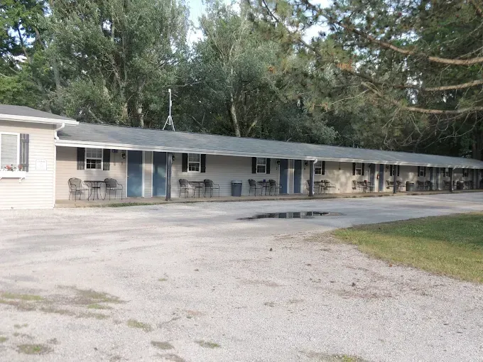 Single-story motel with blue doors and a row of parked chairs along the front, set beside a gravel lot and trees.