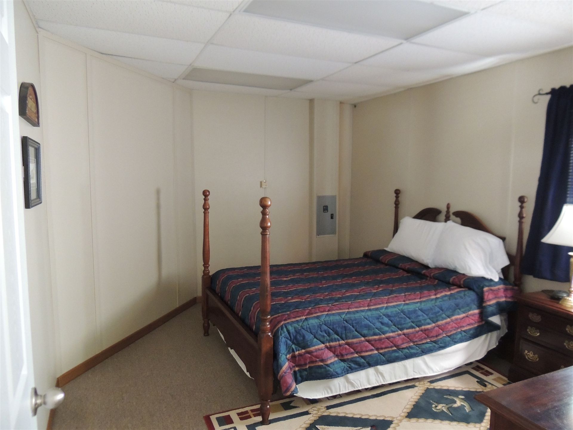 Small bedroom with a wooden bed, blue-and-red quilt, white walls, and a window with dark curtains.