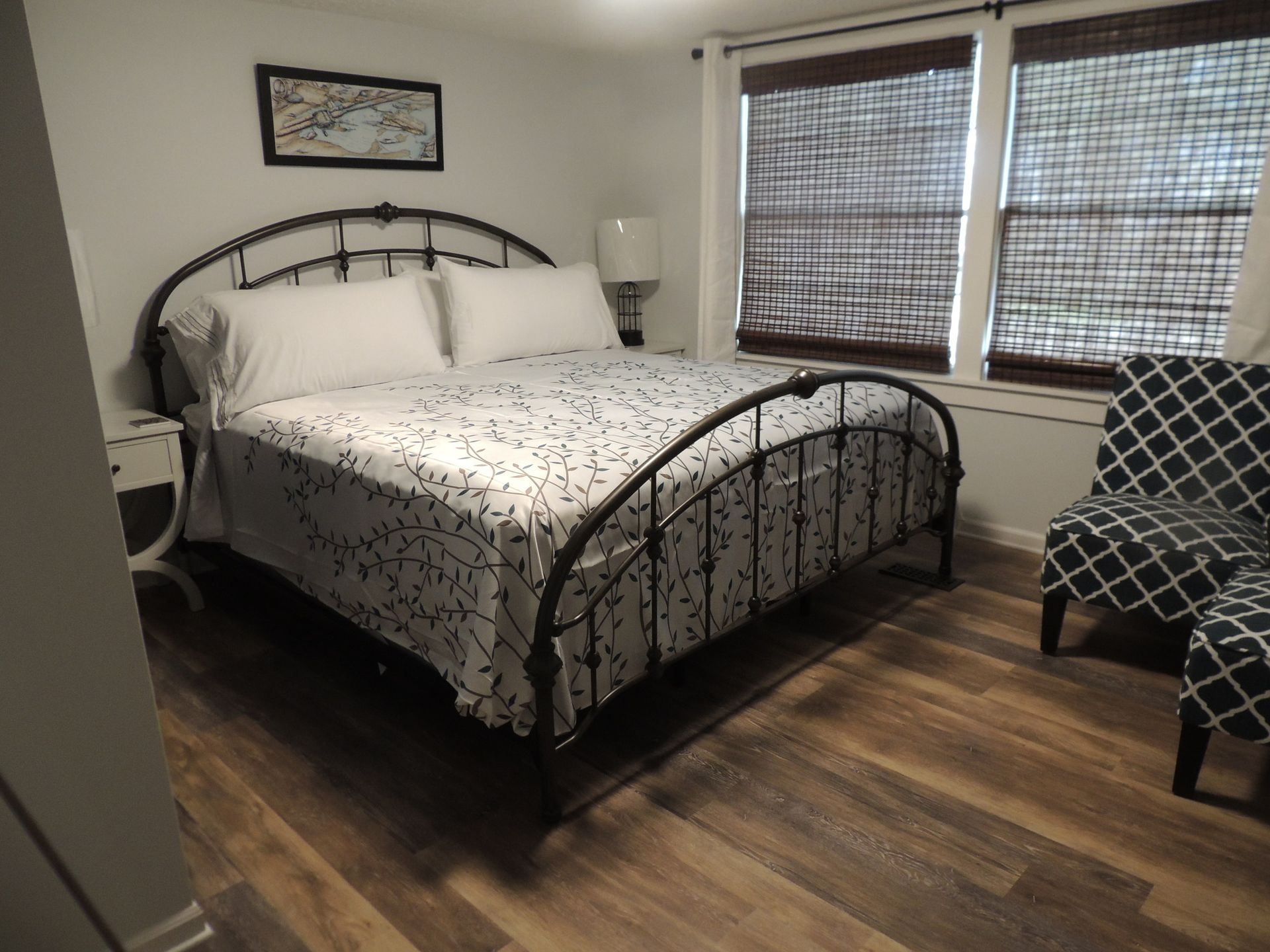 Bedroom with black metal bed, white floral bedding, wood floors, and windows with blinds